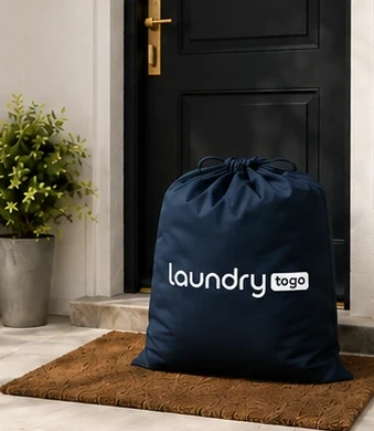 A navy LaundryToGo drawstring bag set on the doormat outside a front door, ready for pickup.